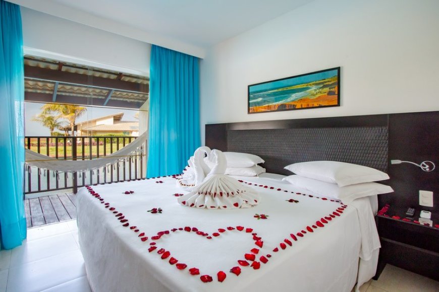 Na imagem, vemos um dos destinos românticos da WAM Experience, uma acomodação no Dom Pedro Laguna. Observamos uma cama com lenço e travesseiros brancos, um detalhe feito com lenço e pétalas de rosas vermelhas. Ainda no quarto, temos, um quadro e, na varada, uma rede.