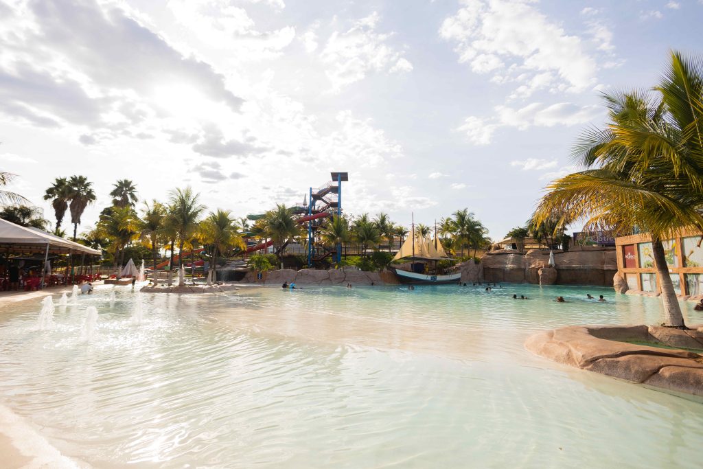 A imagem mostra uma vista panorâmica do parque aquático Náutico Praia Clube em um dia ensolarado. Em primeiro plano, há uma grande piscina com água cristalina e algumas fontes. Ao redor da piscina, há várias palmeiras que dão um aspecto tropical ao ambiente. Ao fundo, é possível ver toboáguas coloridos e uma estrutura em formato de barco, sugerindo um tema náutico. No lado esquerdo da imagem, há uma área coberta com mesas e cadeiras, provavelmente uma área de alimentação. A atmosfera geral é de diversão e relaxamento em um ambiente ao ar livre.