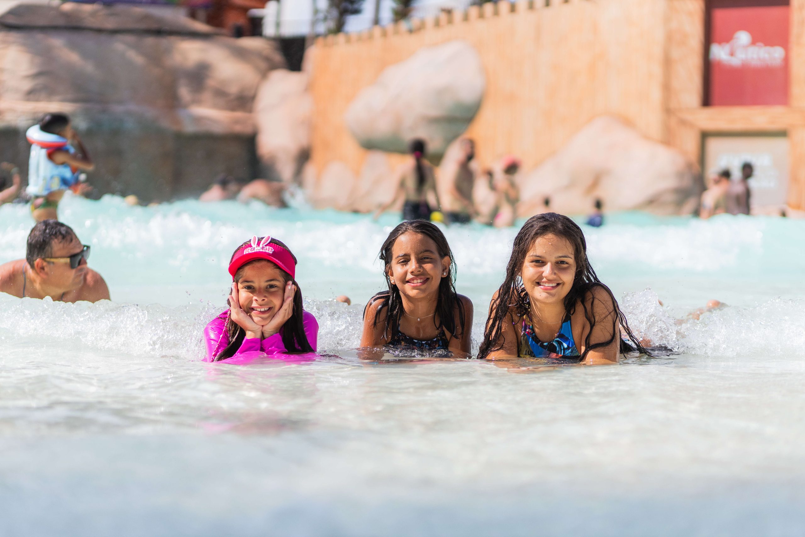 A imagem mostra três meninas deitadas em uma piscina, no Náutico Praia Clube, aproveitando um dia ensolarado. Elas estão sorrindo e parecem estar se divertindo bastante. A menina à esquerda está usando uma camisa de manga longa rosa e um boné vermelho. A menina no meio e a da direita estão usando trajes de banho e ambas têm os cabelos molhados.