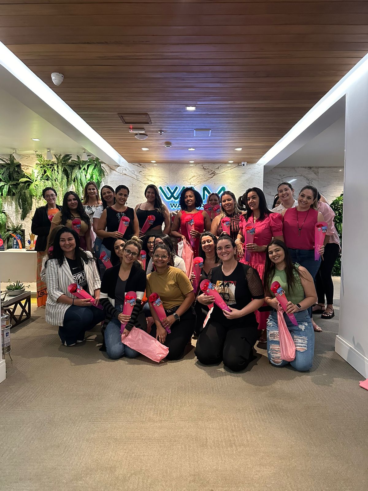 A imagem mostra um grupo de mulheres posando juntas em um ambiente interno. A maioria delas está segurando um objeto rosa em formato de cone. As mulheres estão na área de recepção, com um teto de madeira e uma parede decorada com plantas. Há um logotipo ao fundo com a palavra "WAM Experience", o nome da empresa. As mulheres estão sorrindo e estão vestindo roupas de cores similares, especialmente rosa.