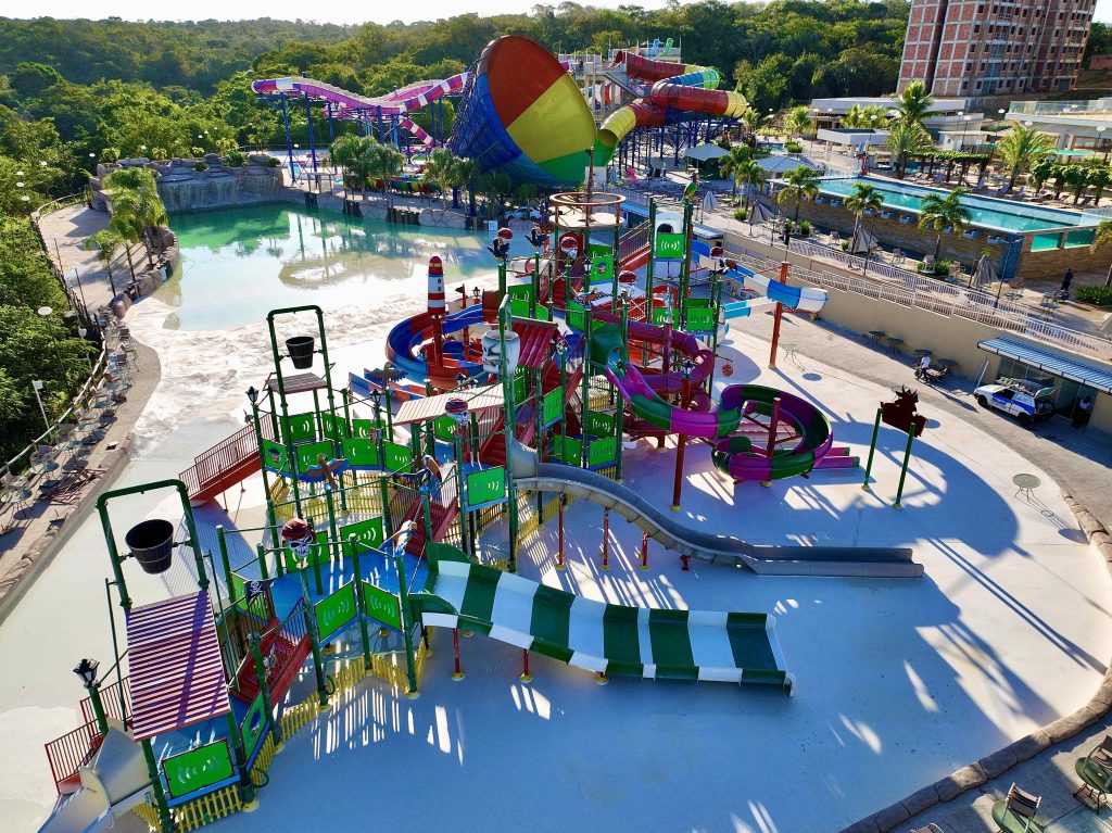 A imagem mostra o Kawana Park com vários escorregadores e uma piscina. Há uma grande piscina azul no fundo da imagem e uma série de escorregadores coloridos, um deles em forma de cone. Na parte inferior da imagem há um escorregador verde e branco, a água está visível em uma parte dele. À direita da imagem há uma área de estar com cadeiras e mesas. No canto superior direito da imagem há um prédio de apartamentos. Há muitos coqueiros por toda a imagem. A reinauguração do Kawana irá atrair muitos visitantes.