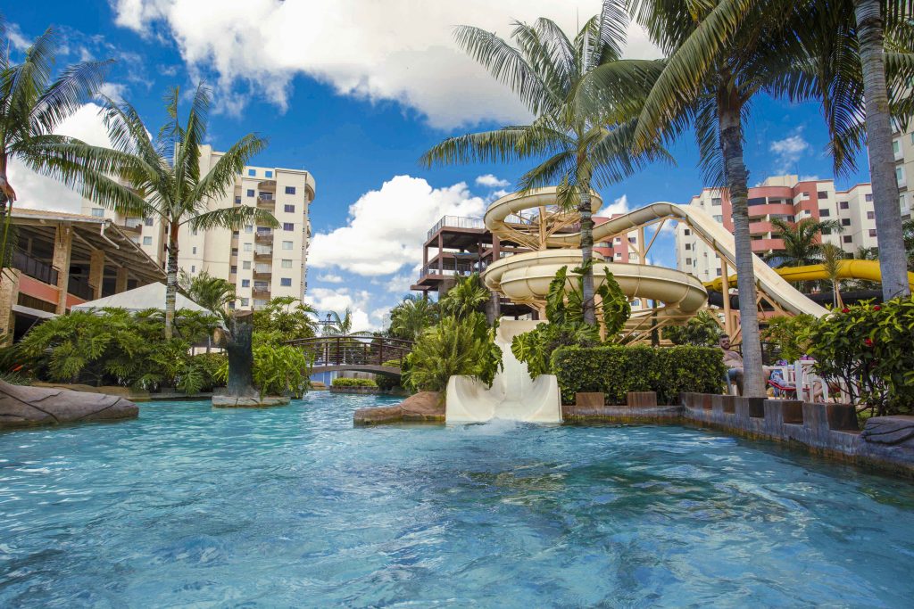 A imagem mostra o Water Park com uma piscina azul brilhante e dois toboáguas brancos e amarelos. Há também um grande tronco de árvore que é parcialmente submerso na água. Na parte de trás do parque aquático, há um prédio alto e uma variedade de vegetação verde. No céu estão nuvens brancas esponjosas.