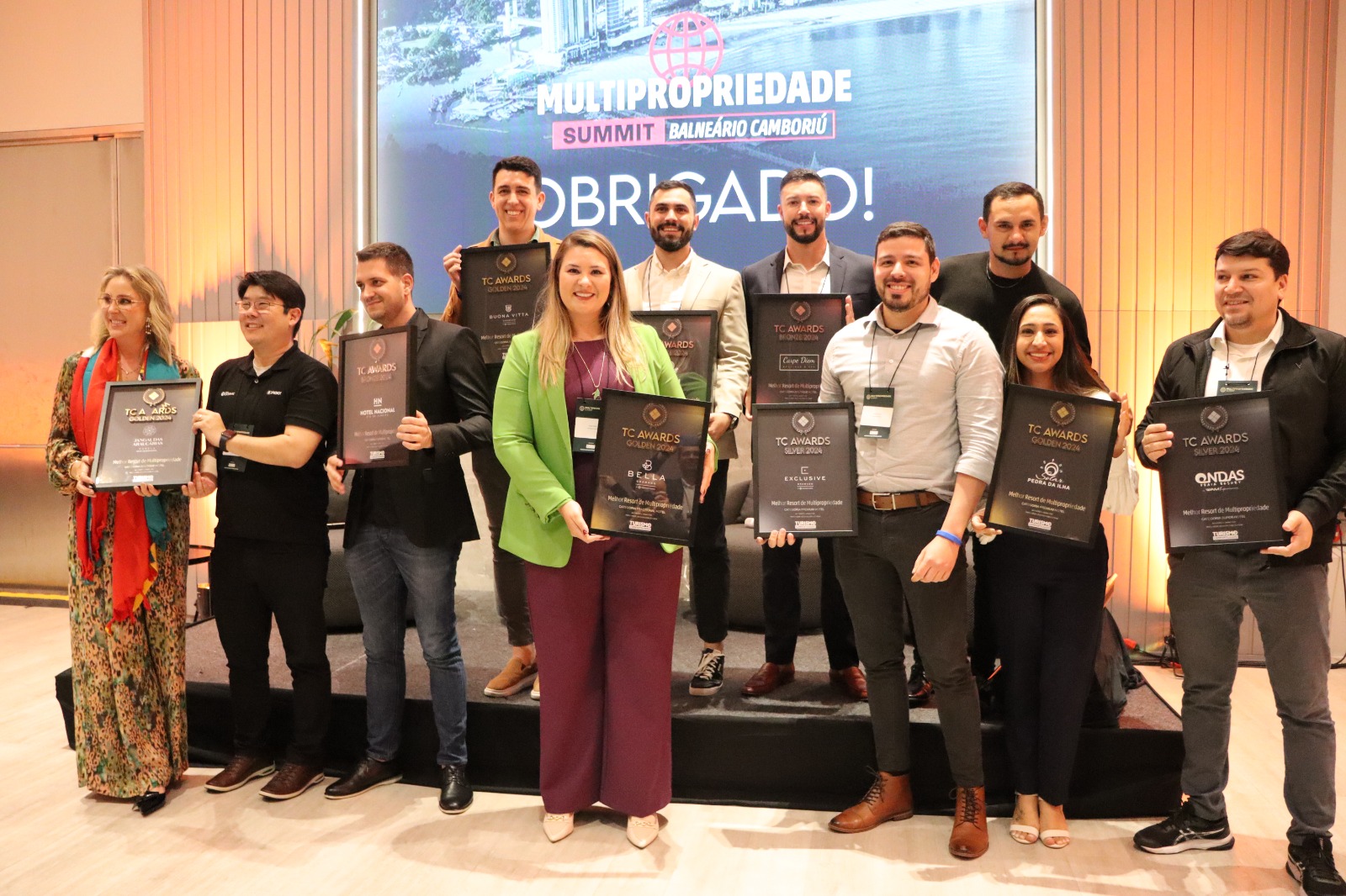 A imagem mostra um grupo de pessoas sorrindo e segurando os prêmios TC Awards 2024. Eles estão em um palco e têm um fundo que diz "Multipropriedade Summit Balneário Camboriu" em português. A placa também diz "Obrigado!" Abaixo das pessoas no palco está uma placa dourada.